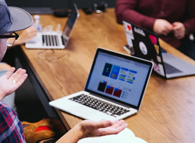 Genrebild på möte mellan personer på en arbetsplats. I förgrunden en man med rutig skjorta och keps på sig med laptop på bordet.