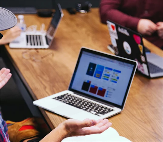 Genrebild på möte mellan personer på en arbetsplats. I förgrunden en man med rutig skjorta och keps på sig med laptop på bordet.